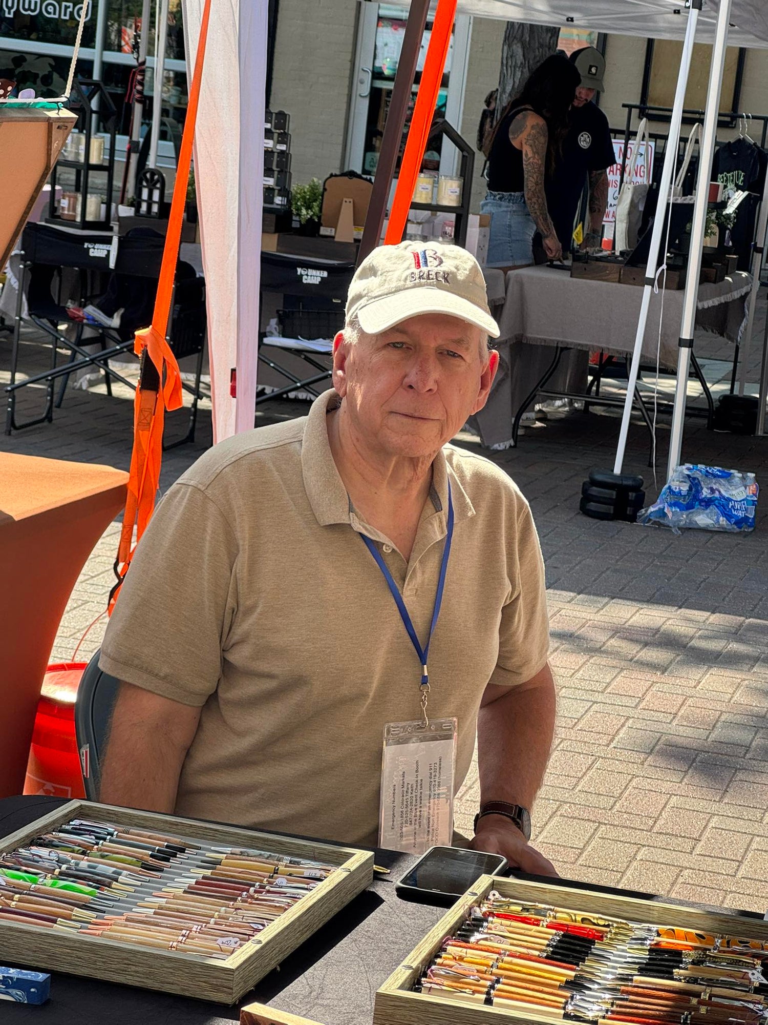 Man with a cap and lanyard standing behind a table with art supplies at an outdoor event.